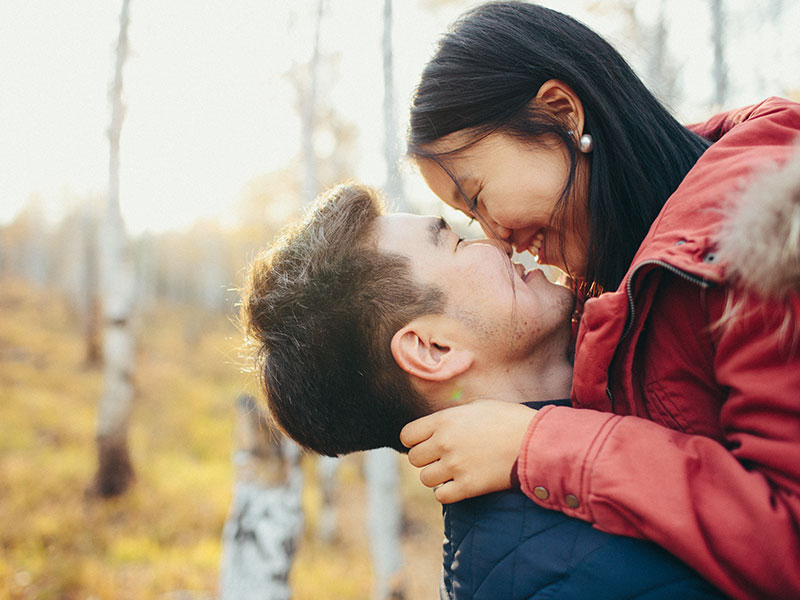 A couple who's best friends with each other smiling and laughing as they kiss in the woods.